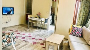 Una sala de estar con un sofá, una mesa de centro, un televisor, una zona de comedor con una mesa y sillas, y una cocina visible a través de una puerta abierta.