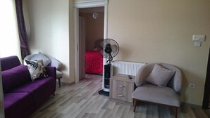 Una sala pequeña con un sofá morado, un sillón gris y un ventilador de pie. Se ve una cama roja al fondo y un radiador en el suelo.