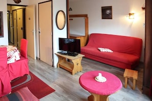 A room with a red sofa, a television on a wooden stand, a small round table with a white object, and a red chair.