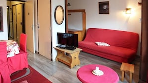 Een kamer met een rode bank, een televisie op een houten dressoir, een klein rond tafeltje met een wit object, en een rode stoel.
