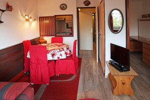 A room with a dining table, chairs, a TV on a wooden stand, a mirror, and a door leading to another room.