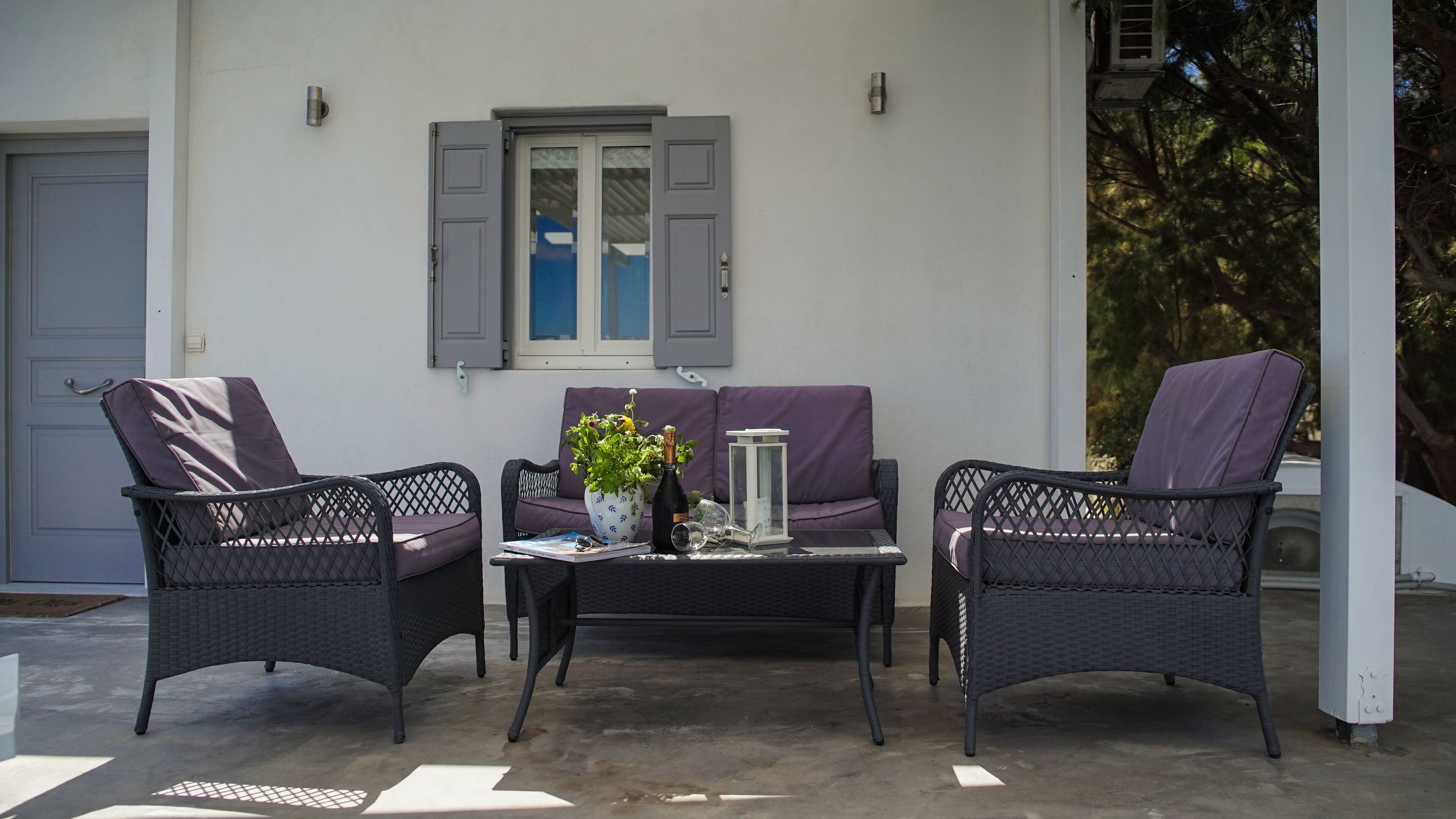 An outdoor seating area with wicker chairs and a sofa, a table with a bottle and glasses, and a window with shutters.
