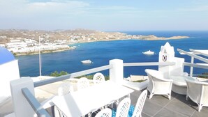 A terrace with white furniture overlooking a coastal view with boats and a distant town. - Grand Panoramic Villa Ozar (Mykonos)