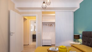 Quarto moderno com sofá amarelo, uma pequena mesa com bule e prato amarelos, poltrona cinza, armário com roupas penduradas e uma parede de destaque azul-esverdeado.