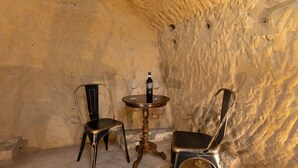 Um quarto pequeno e rústico com parede de pedra, uma mesa de madeira redonda, duas cadeiras de metal e uma garrafa de vinho.