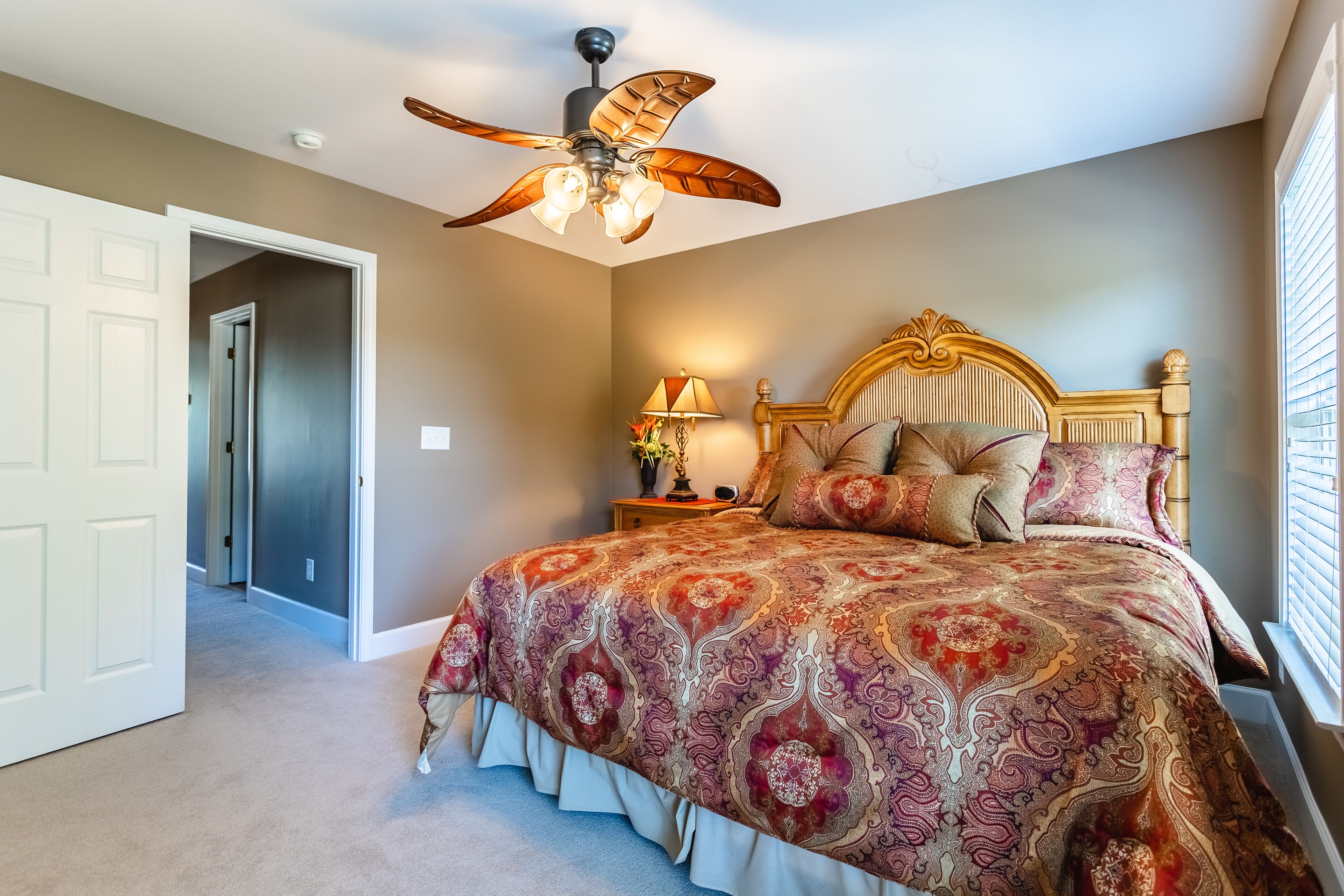 A bedroom with a large bed, a ceiling fan, a nightstand with a lamp, and a window with blinds.