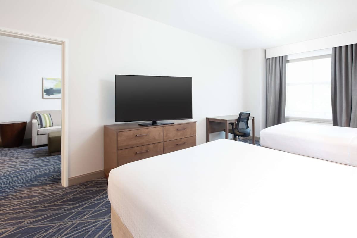A hotel room with a large bed, a flat-screen TV on a wooden dresser, a desk with a chair, and a window with curtains.