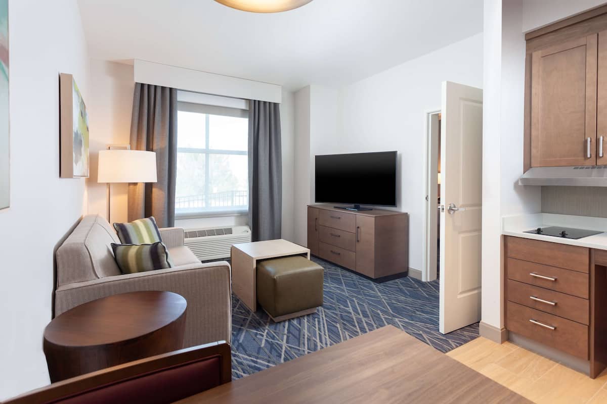 A hotel room with a sofa, ottoman, flat-screen TV, kitchenette, and a large window with curtains.