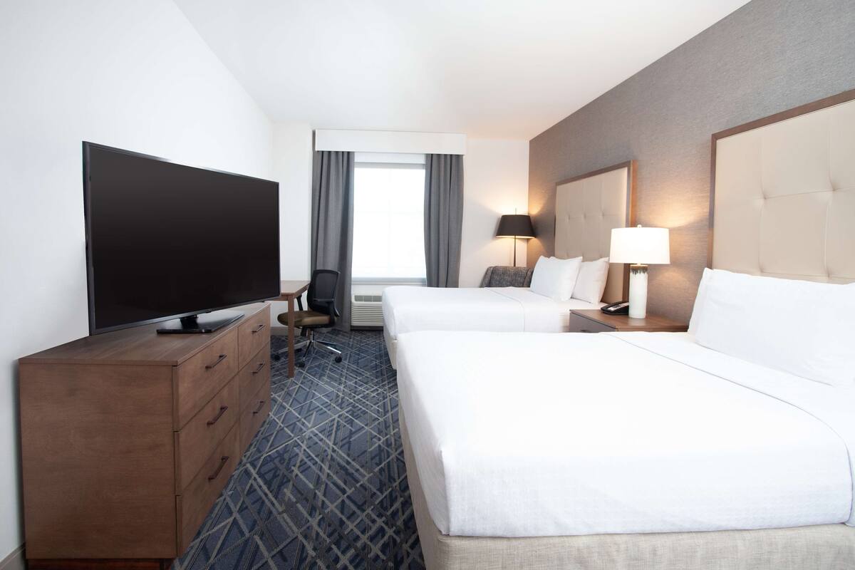A hotel room with a flat-screen TV, two beds, a desk, a chair, and a window with curtains.