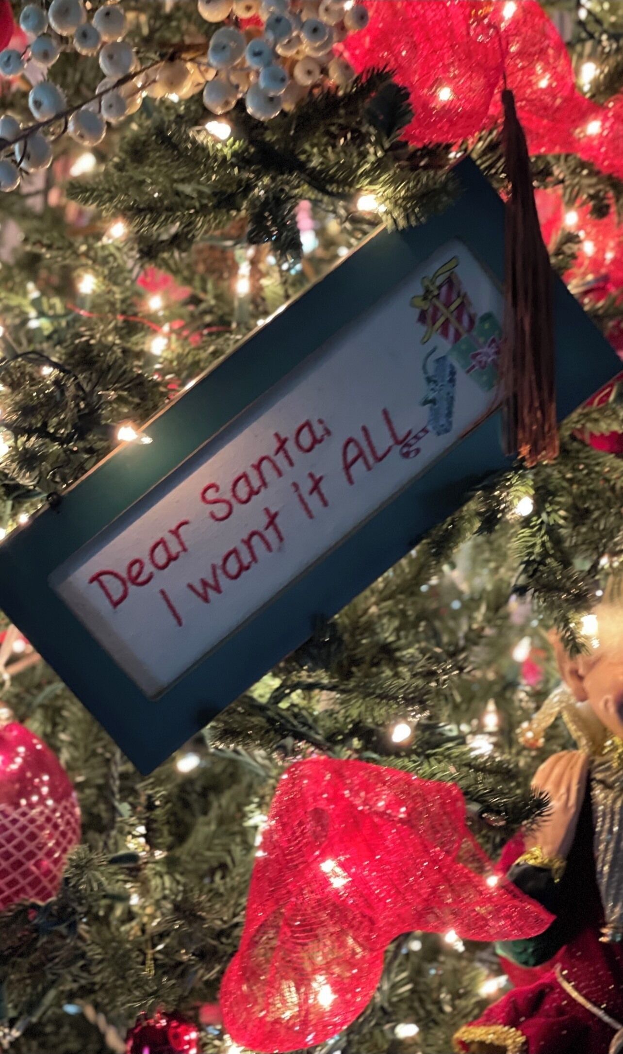 Un sapin de Noël avec une pancarte sur laquelle on peut lire : « Cher Père Noël, je veux tout. »