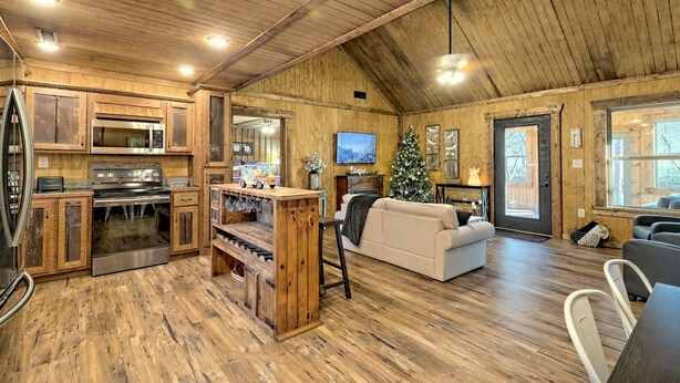A rustic cabin interior with wooden walls, a kitchen with a stove and microwave, a living area with a couch and TV, and a Christmas tree.