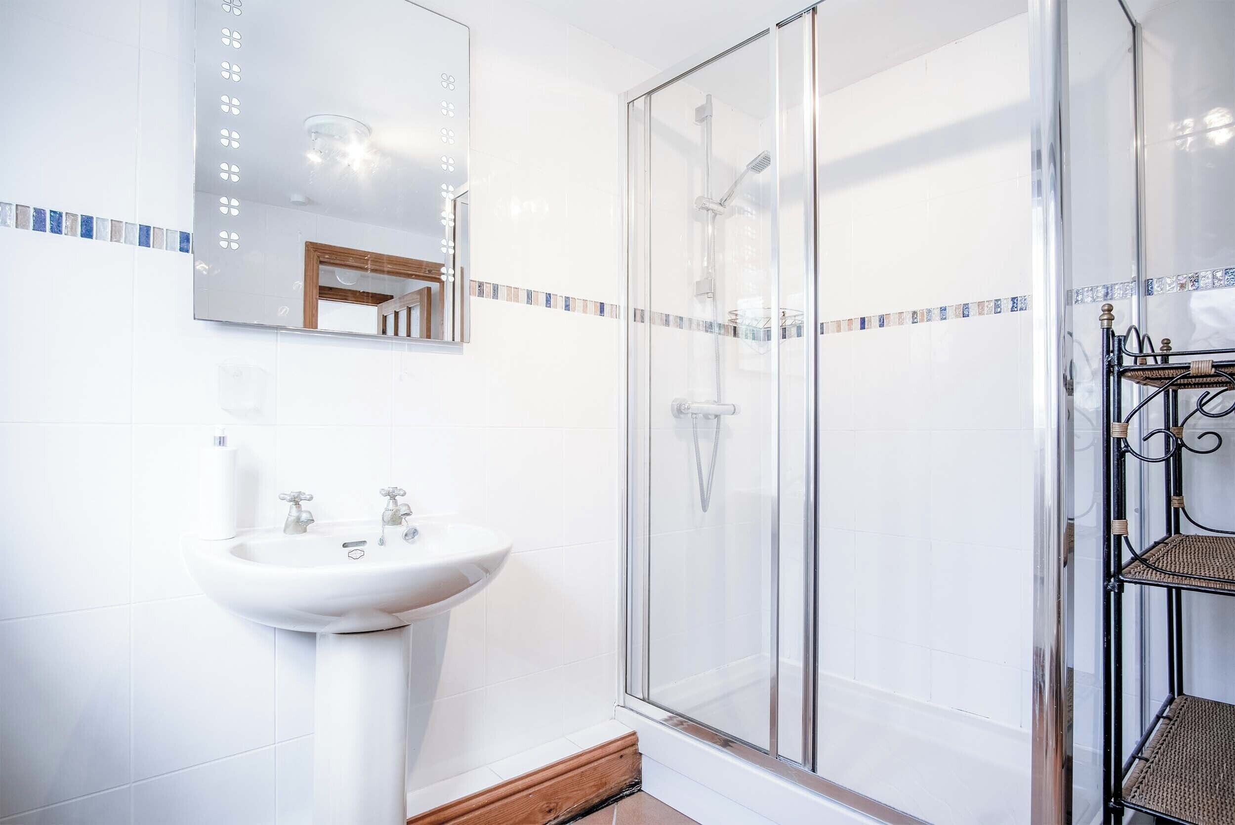 Een badkamer met een glazen douchecabine, een witte wastafel en een spiegel met een decoratieve tegeltjesrand.