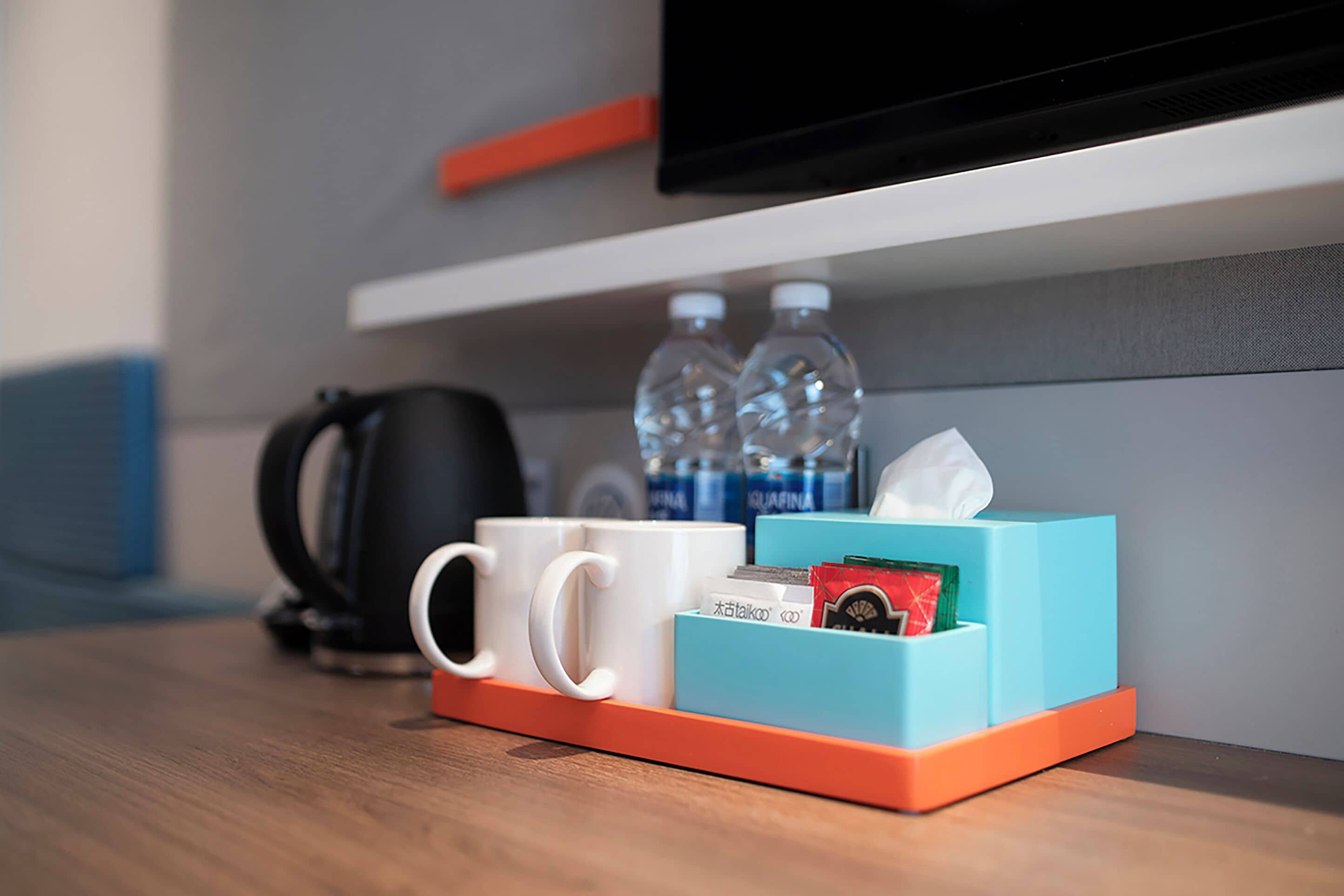 A hotel room with a wooden table, a coffee maker, two water bottles, a tissue box, and a remote control.