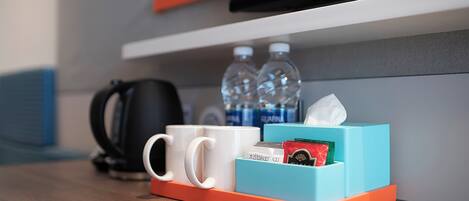 A hotel room with a wooden table, a coffee maker, two water bottles, a tissue box, and a remote control.