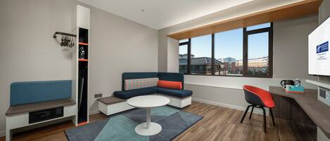 A modern hotel room with a seating area, a small table, and a view of the city through large windows.