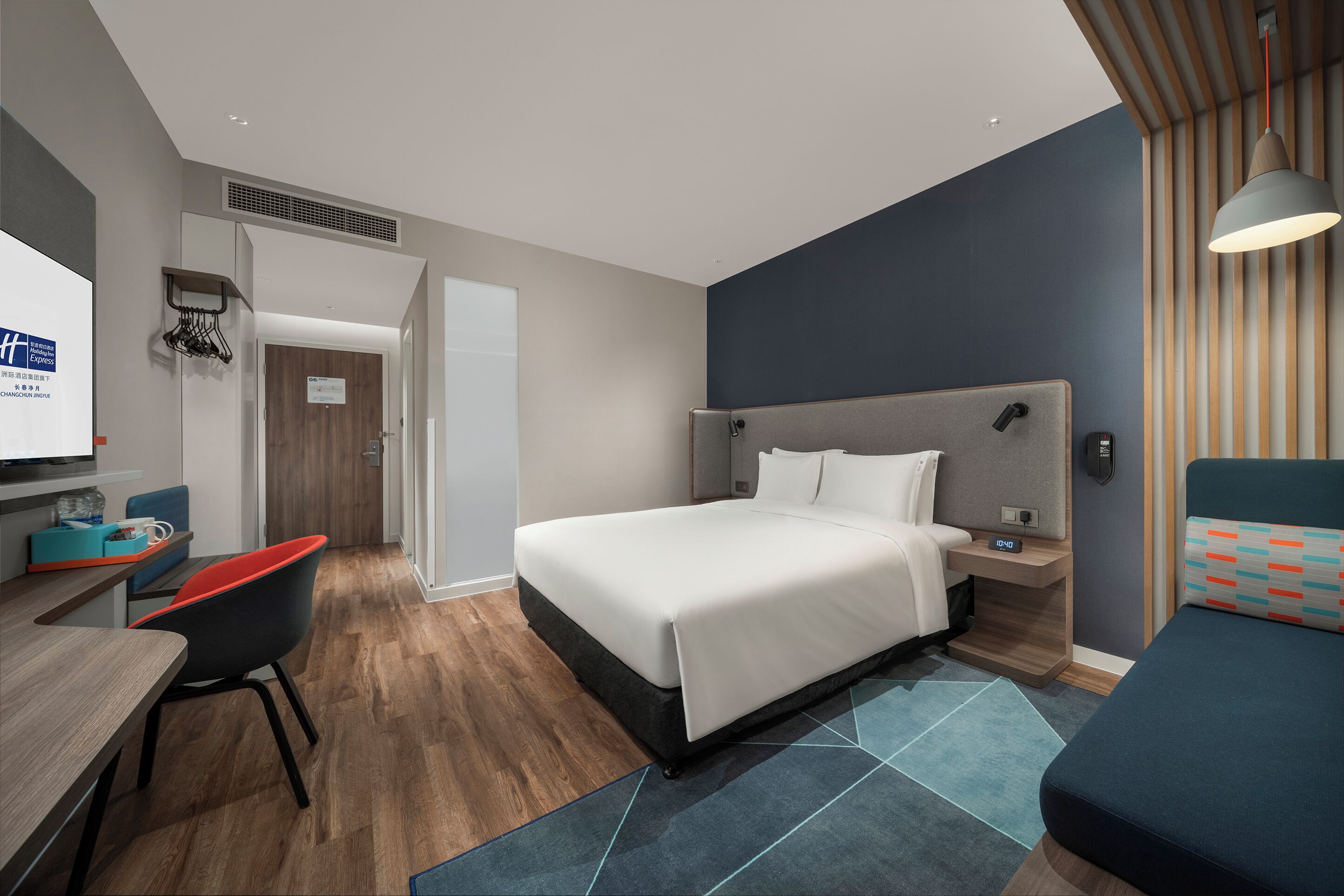A modern hotel room with a bed, a desk with a chair, a bedside table with a lamp, and a view of the hallway.