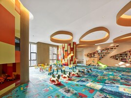 Un espacio interior bien iluminado con diseños geométricos coloridos en las paredes, una zona de juegos con juguetes y un suelo vistoso, además de una escalera con estanterías integradas.