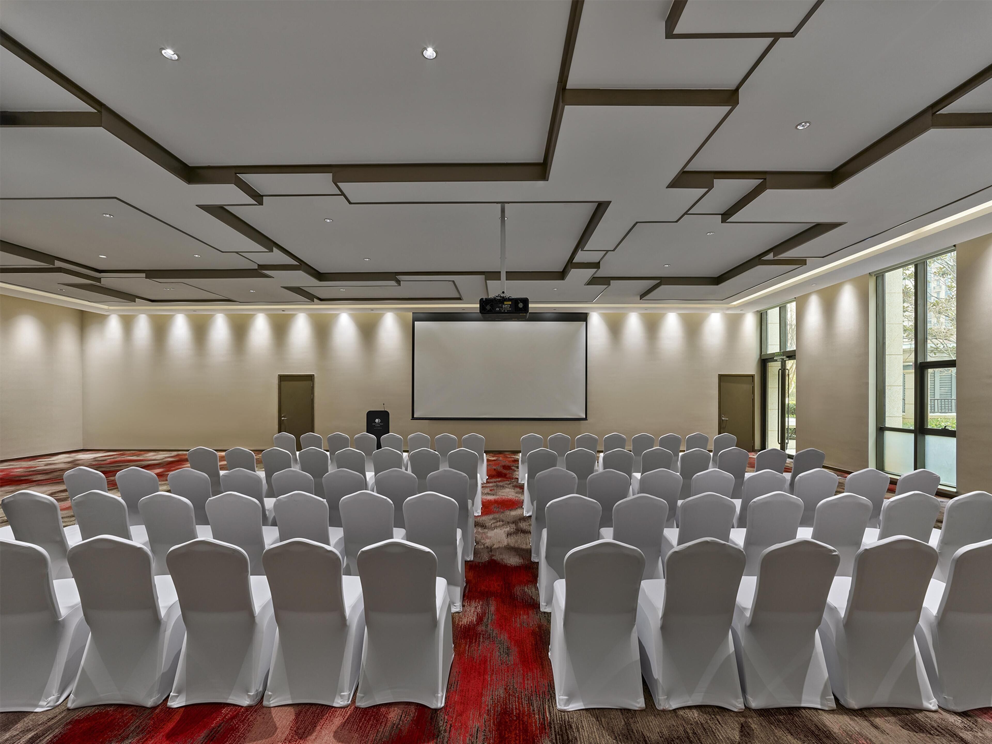 Un salón de conferencias con hileras de sillas blancas, una pantalla grande y un proyector.