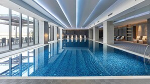 Una piscina interior moderna con paredes de vidrio transparente, suelo de baldosas e iluminación lineal en el techo.