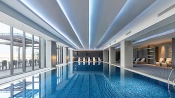 A modern indoor swimming pool with clear glass walls, a tiled floor, and a ceiling with linear lighting.