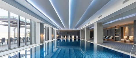 A modern indoor swimming pool with clear glass walls, a tiled floor, and a ceiling with linear lighting.