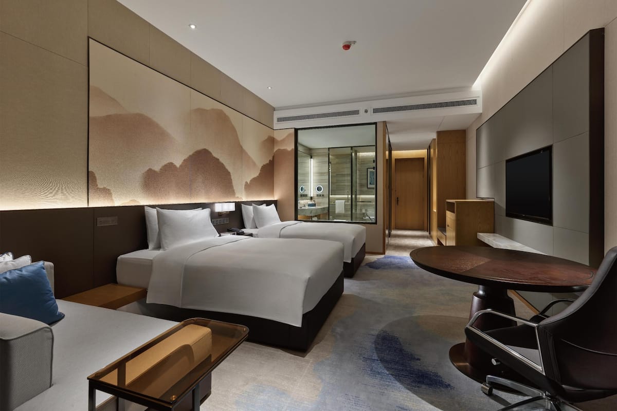 Ein modernes Hotelzimmer mit einem großen Bett, einem Schreibtisch, einem Stuhl, einem Fernseher und einem verglasten Badezimmer.