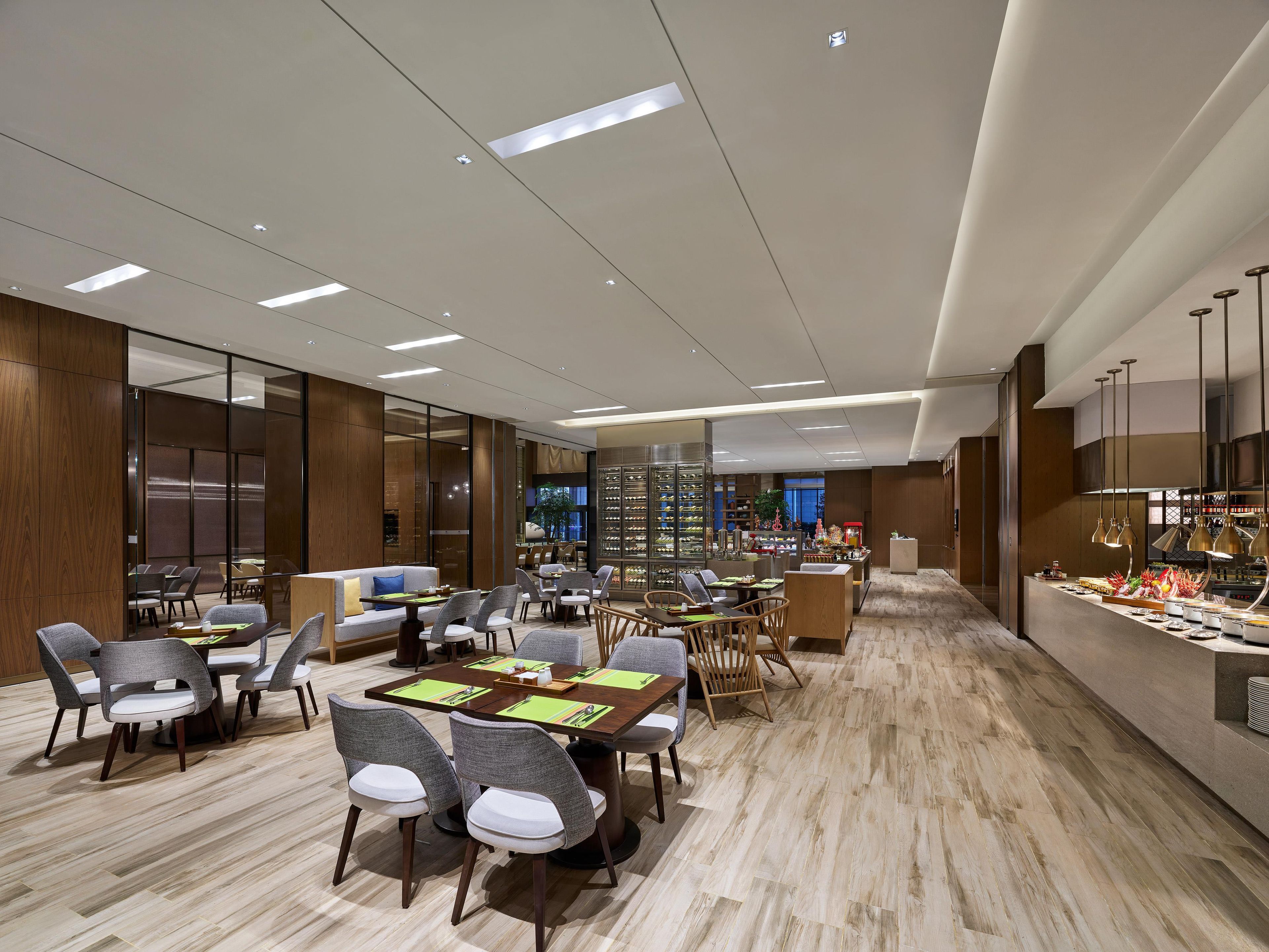 Un área de comedor de hotel moderna con varias mesas y sillas, un bar central, y vista a la cocina al fondo.