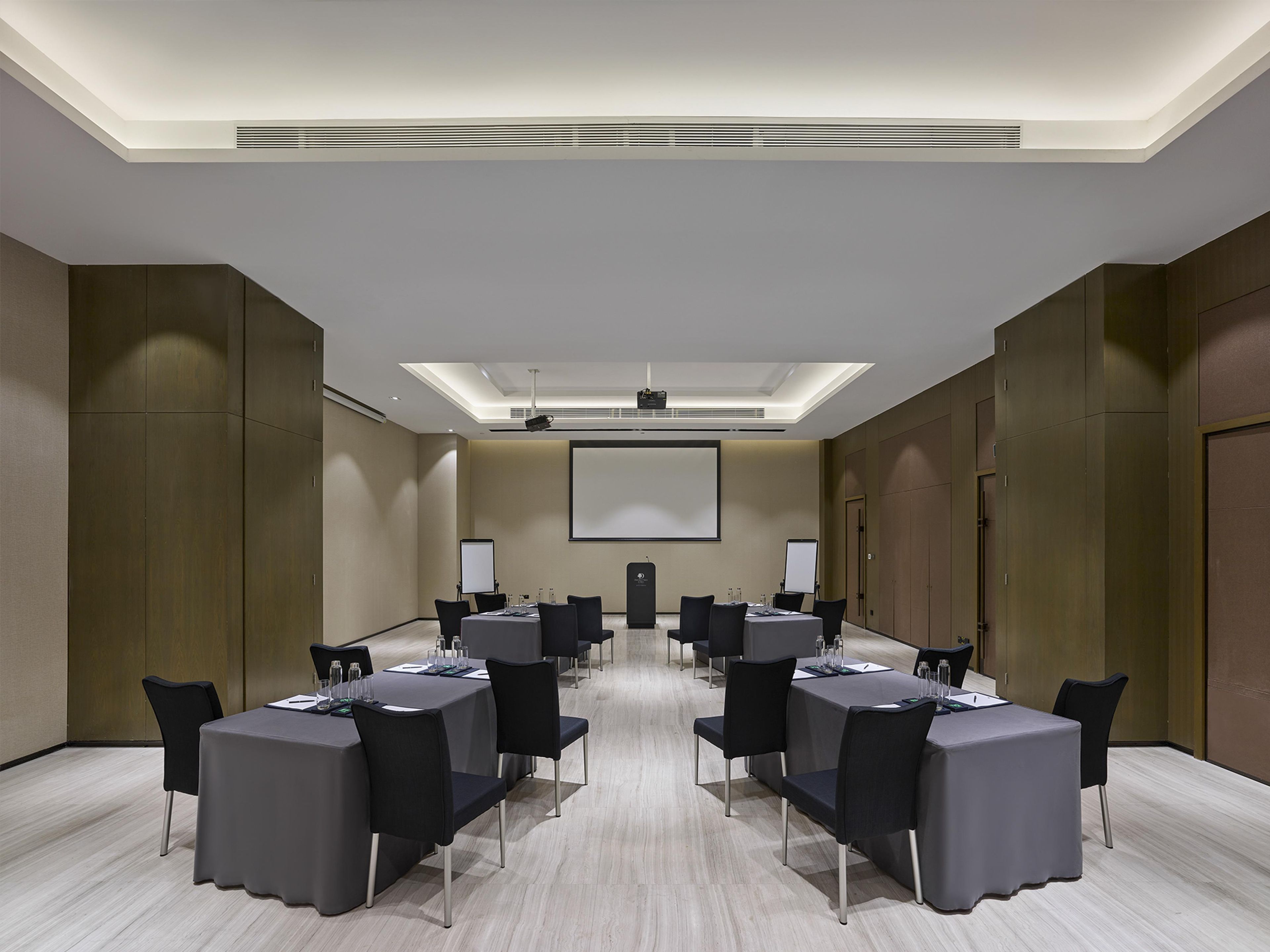 Une salle de conférence dotée d’un écran de projection central, de plusieurs tables avec des chaises et d’un plafond équipé d’un éclairage encastré.