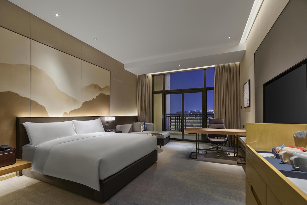 Ein modernes Hotelzimmer mit einem großen Bett, einem Schreibtisch, einem Stuhl, einem Computer und Blick auf die nächtliche Skyline.