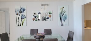 Uma sala de jantar com mesa de vidro, seis cadeiras e três quadros florais na parede.