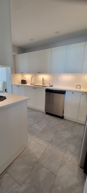 Uma cozinha moderna com armários brancos, uma lava-louças de aço inoxidável e uma bancada com uma chaleira e um rolo de papel toalha.