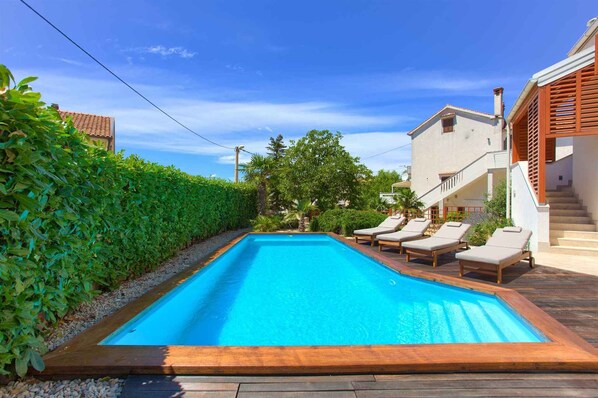 A rectangular swimming pool with lounge chairs, surrounded by a wooden deck and a hedge.