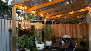 An outdoor patio with a wooden pergola, string lights, a grill, and potted plants.