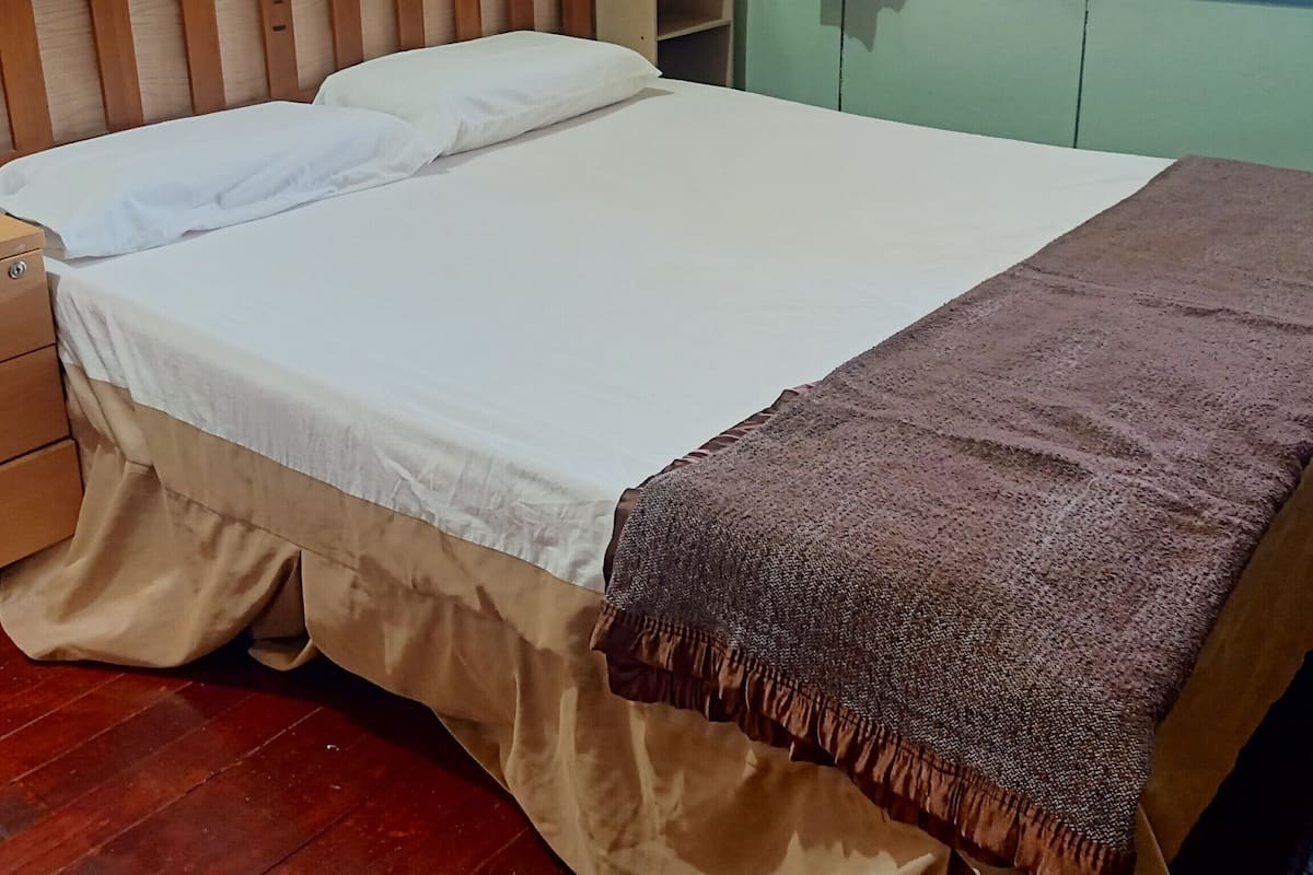 Une chambre avec un lit simple, une tête de lit en bois, une table de chevet, un rideau vert et une fenêtre donnant sur l’extérieur.