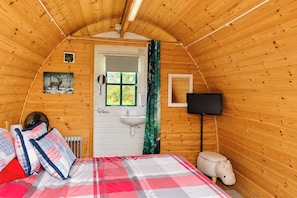 A cozy cabin bedroom with a bed, a bedside table, a mirror, a window with a view, a television, and a radiator.