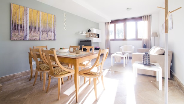 Un comedor con una mesa y sillas de madera, una cocina al fondo y una sala de estar con un sofá y una lámpara.