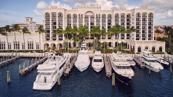 A marina with multiple yachts docked in front of a large, multi-story hotel with arched windows and balconies.