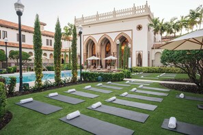 A courtyard with yoga mats arranged on artificial grass, a pool, and a building with arched windows and a decorative facade.