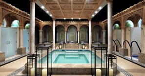 A luxurious indoor pool area with ornate arches and a central pool, surrounded by columns and candle-lit tables.