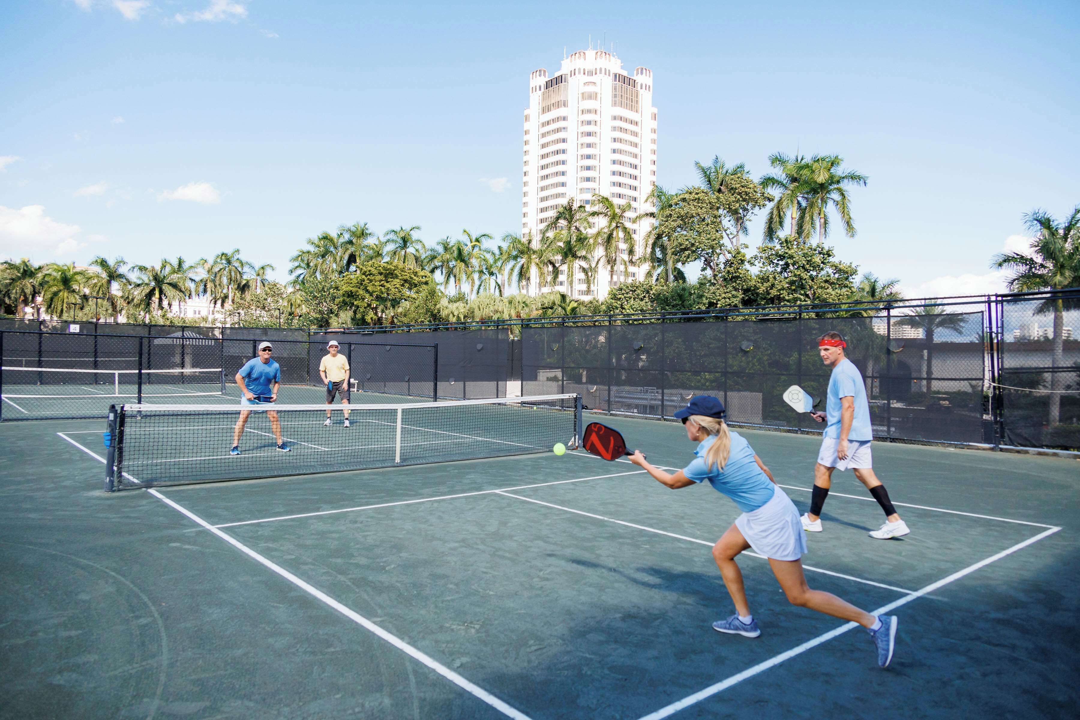 A tennis court with a net, palm trees, and a tall building in the background.