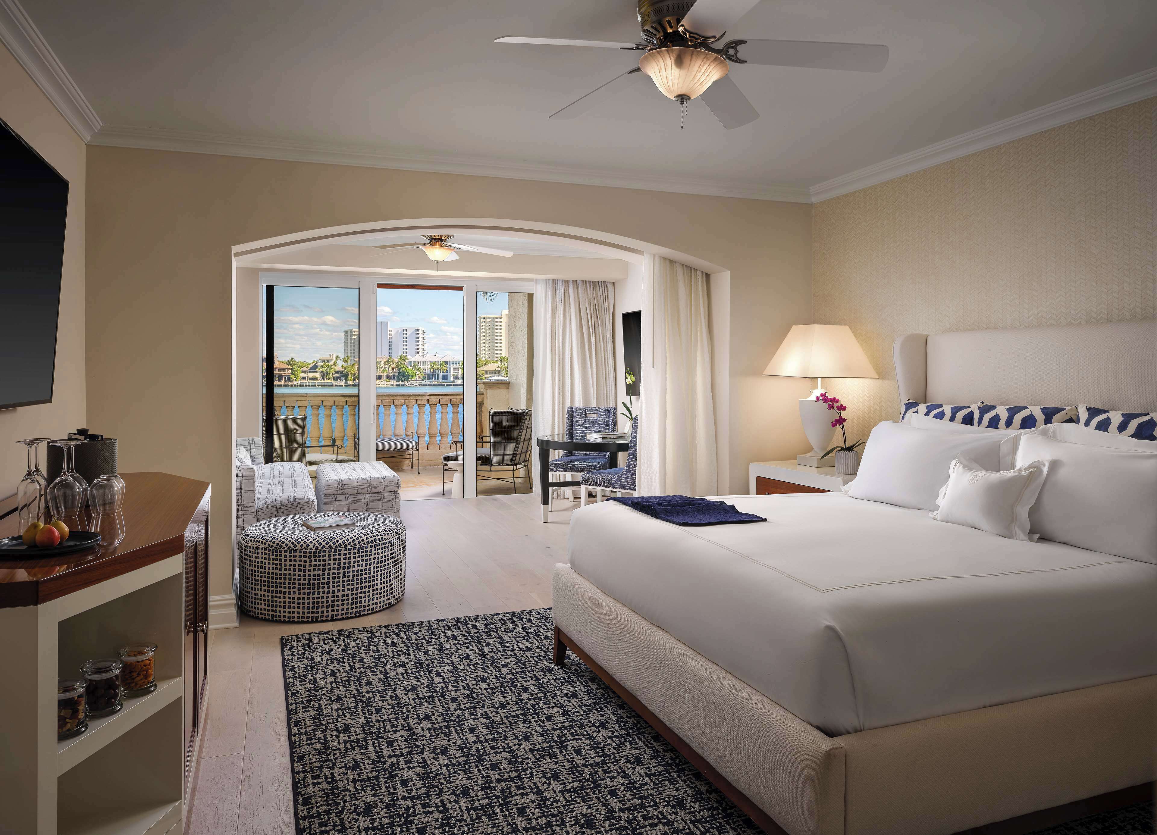 A hotel room with a large bed, a desk with a lamp, a chair, a small table with a vase, and a view of the city through a balcony.