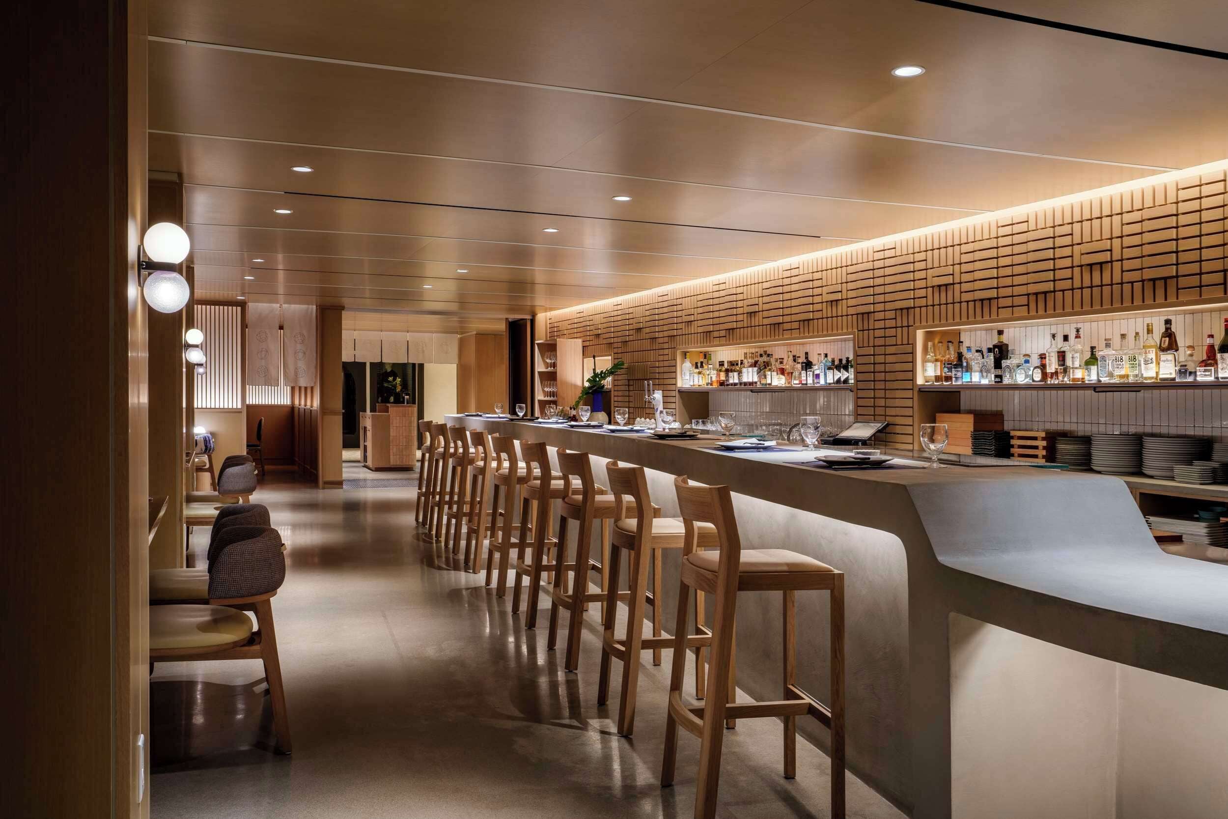 A modern bar area with a long counter, wooden bar stools, and a variety of bottles and glasses.