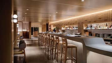 A modern bar area with a long counter, wooden bar stools, and a variety of bottles and glasses.