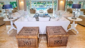 Ein luxuriöses Wohnzimmer mit weißen Sofas, einem Couchtisch und Korbwannen, auf denen ‘1953’ steht. Im Hintergrund sind durch die Glastüren ein Pool sichtbar.