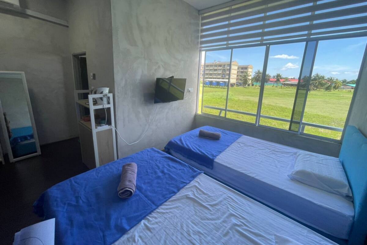 A hotel room with two beds, a TV on the wall, and a view of a grassy field and buildings outside.