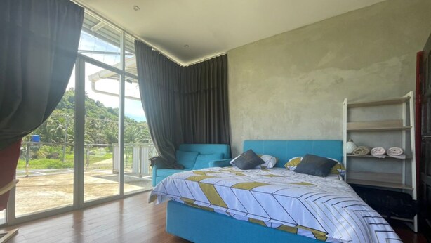 A bedroom with a large bed, a blue headboard, a window with curtains, and a view of greenery outside. - OYO Home 90370 D'tampi Homestay (Semporna)