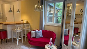 A compact living space with a red sofa, a small round table with a vase, a kitchen area with a wooden countertop, and a large mirror reflecting the room. - Spacious room for two (Dömitz OT Rüterberg)