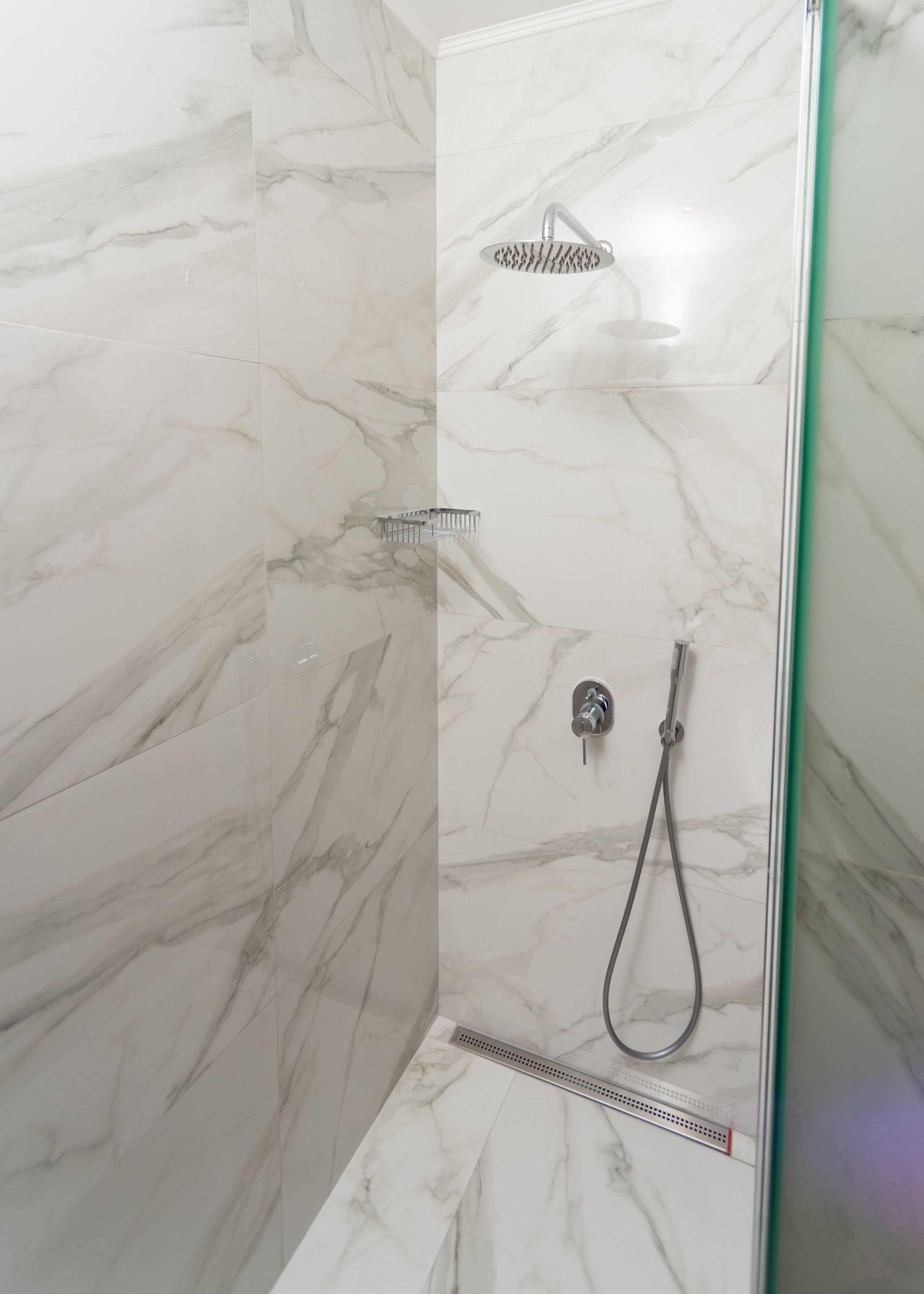 A modern shower with a handheld showerhead and a wall-mounted faucet, set in a room with marble walls and floor.