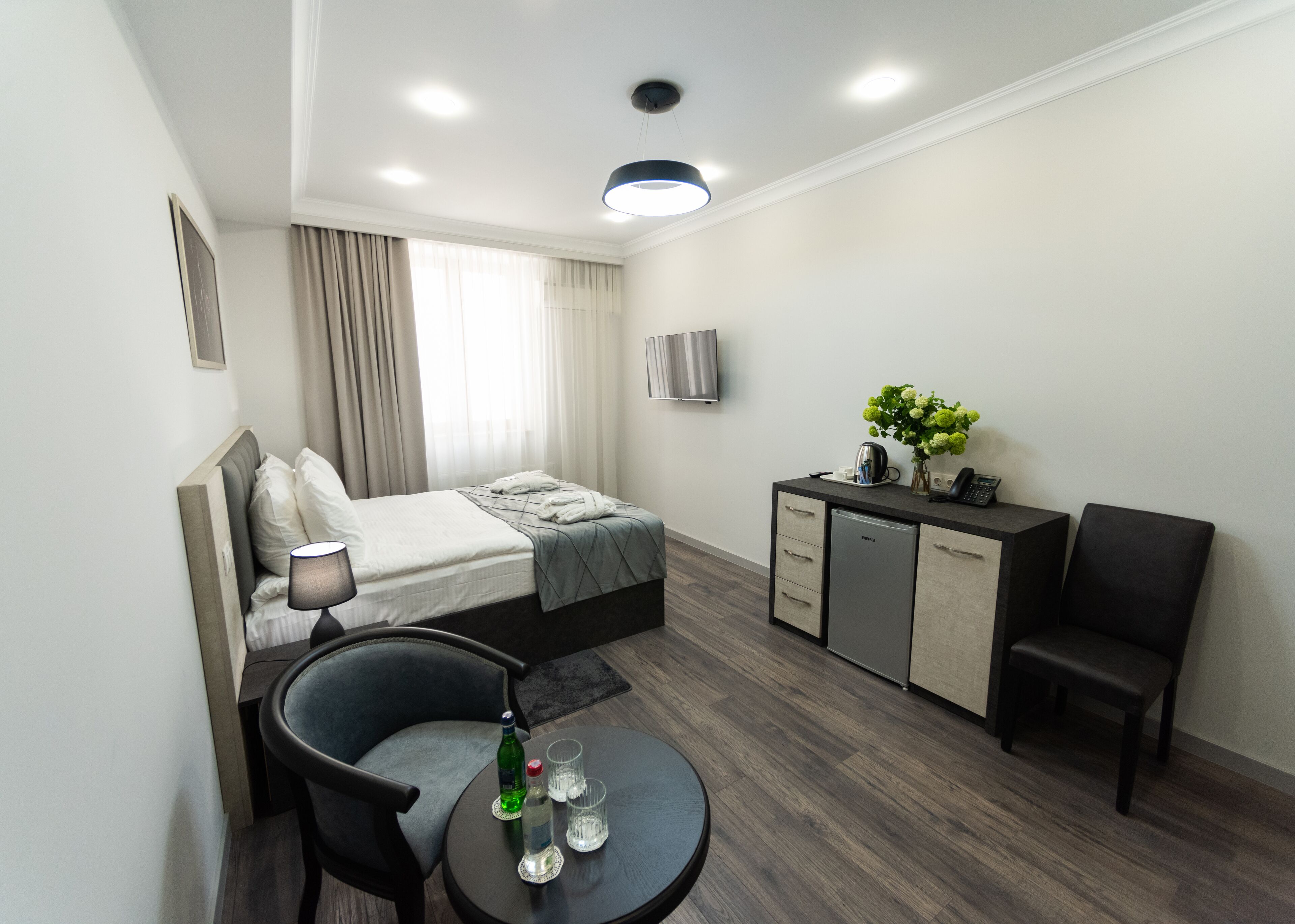 A modern hotel room with a bed, a desk, a chair, a small table with glasses and a bottle, and a kitchenette.