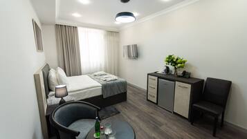 A modern hotel room with a bed, a desk, a chair, a small table with glasses and a bottle, and a kitchenette.